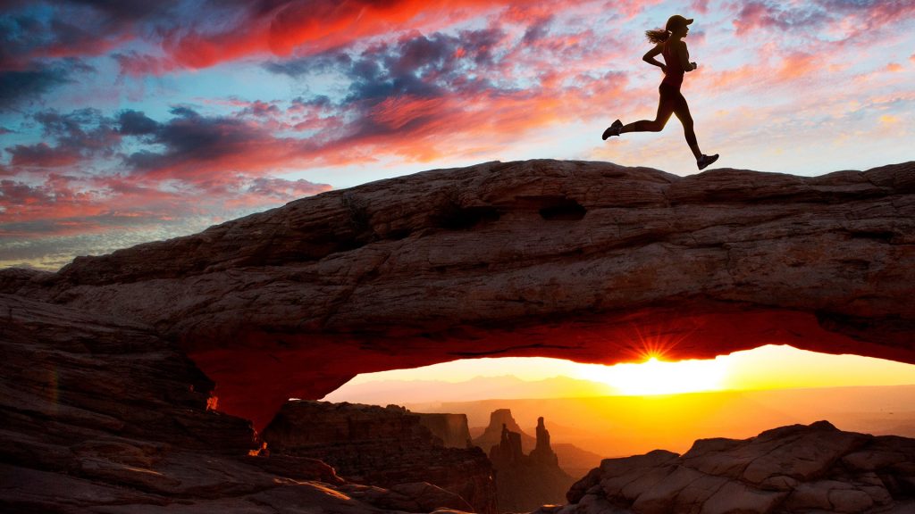 Young female runner running over Mesa Arch rock formation at sunset, Moab, Utah, USA