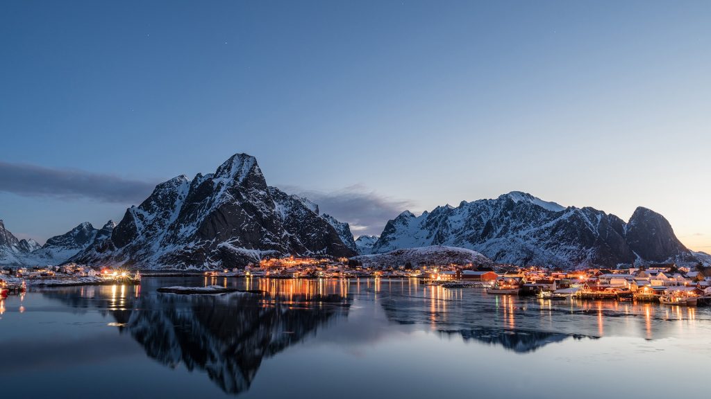 Village illumination with mountain range reflection on coastline at dawn, Lofoten Islands, Norway